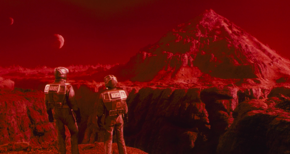 <p><strong>Figure 1.1</strong> The first appearance of the Pyramid Mines logo, seen on the portable life support units worn by Quaid and Melina, as they stand before a towering pyramid-like mountain on Mars’ surface. The logo’s triangular form a simplified representation of this mountain, within which the mines are operated.</p>